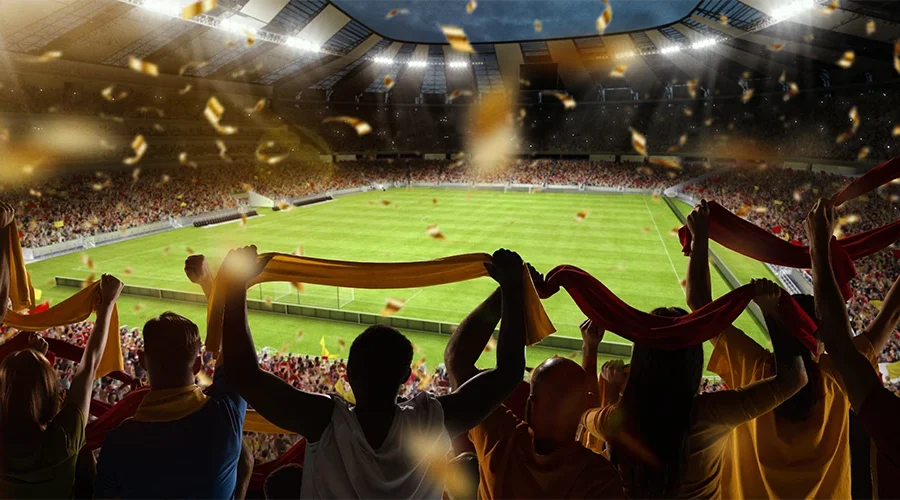 Ein lebhaftes Stadionbild während eines Fußballspiels. Im Vordergrund jubeln zahlreiche Fans in verschiedenfarbigen Trikots, einige mit Schals und Flaggen in den Händen. Im Hintergrund ist das grüne Spielfeld sichtbar, umgeben von weiteren Zuschauertribünen. Die Atmosphäre ist energiegeladen und festlich.