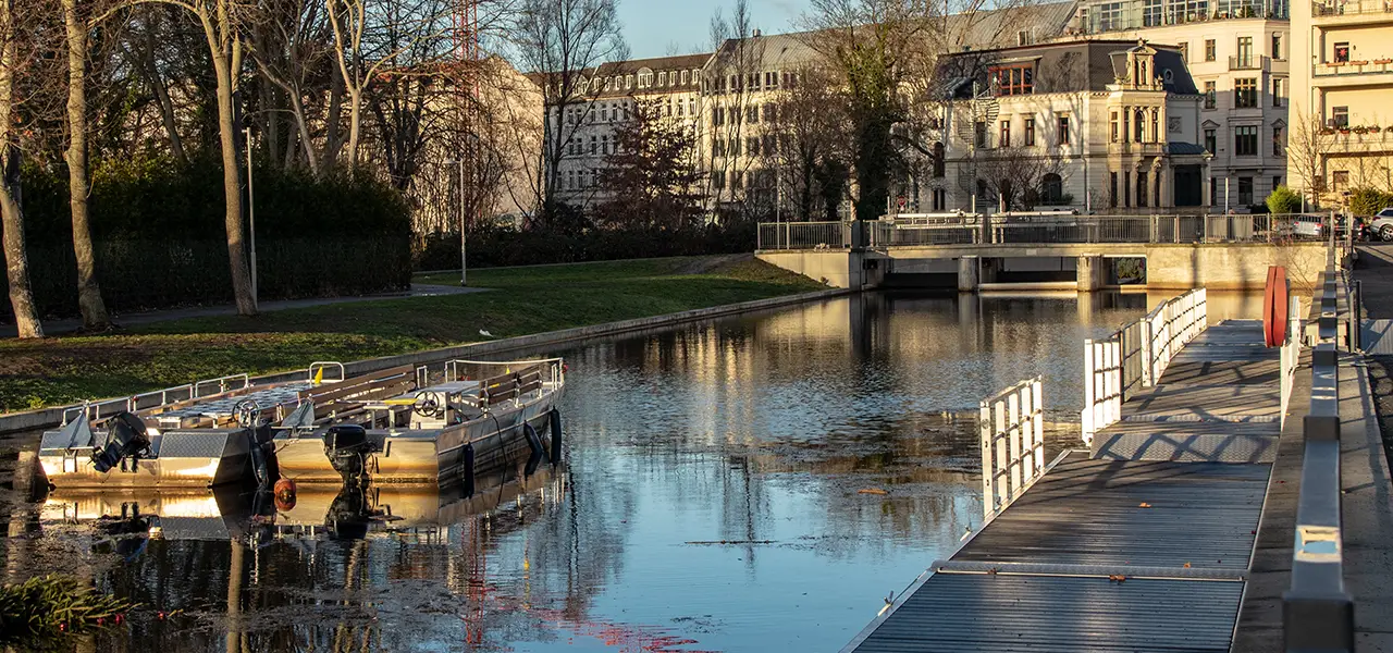 Zwei leere Motorboote liegen an einem Steg am stadthafen Leipzigauf einem ruhigen Kanal, umgeben von Bäumen und historischen Gebäuden im Hintergrund. Ein Baukran ragt über die Szenerie hinaus.