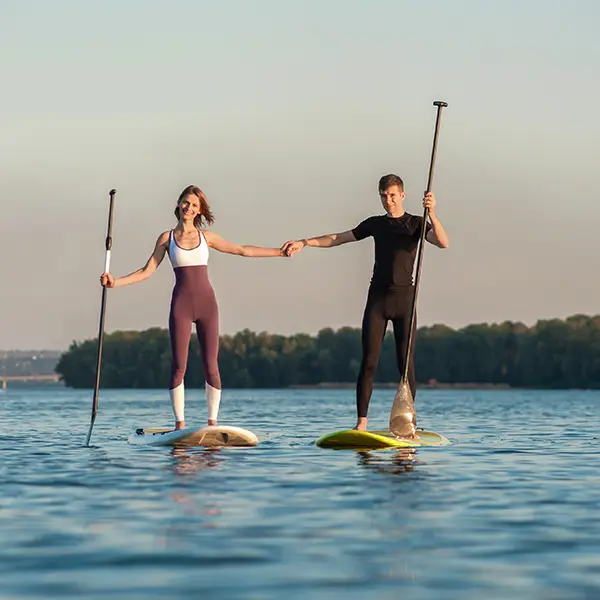 Ein Mann und eine Frau stehen auf Paddleboards auf einem ruhigen See und halten sich an den Händen.