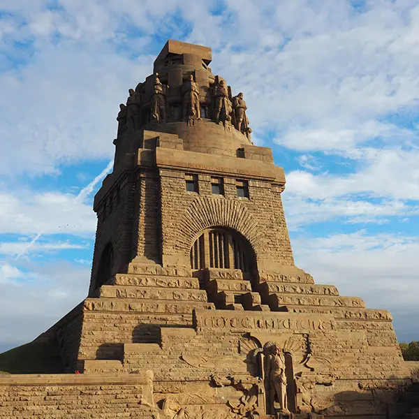 Frontalansicht des Völkerschlachtdenkmals in Leipzig bei sonnigem Wetter, das massive steinerne Bauwerk ragt in den blauen Himmel.