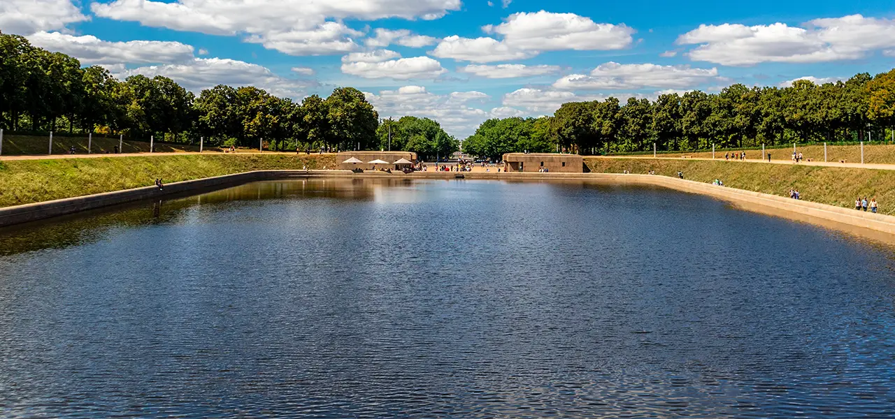 Blick auf das Wasserbecken vor dem Völkerschlachtdenkmal in Leipzig, umgeben von Bäumen und Spazierwegen bei sonnigem Wetter.