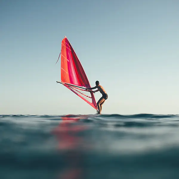 Eine Person auf einem Windsurfbrett mit rotem Segel auf offenem Wasser bei ruhiger See und klarem Himmel.