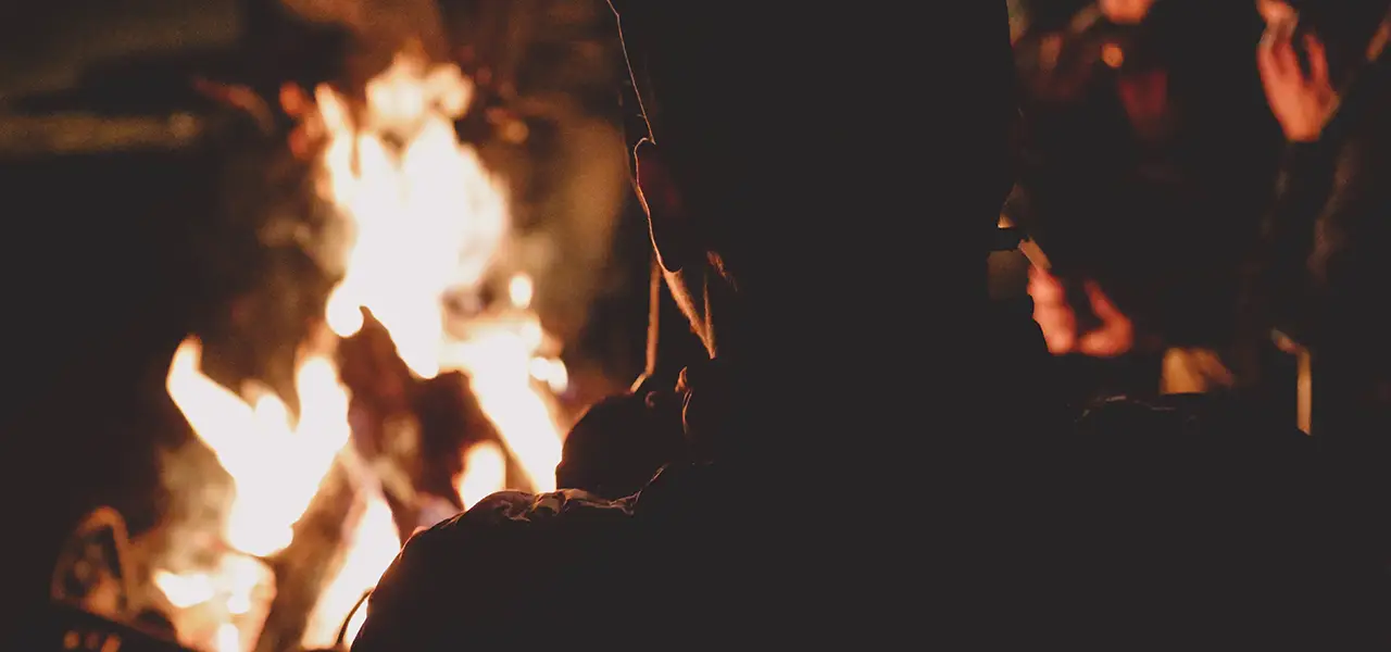Eine Gruppe von Menschen sitzt um ein loderndes Lagerfeuer, das warme Licht erhellt ihre Silhouetten in der Dunkelheit.