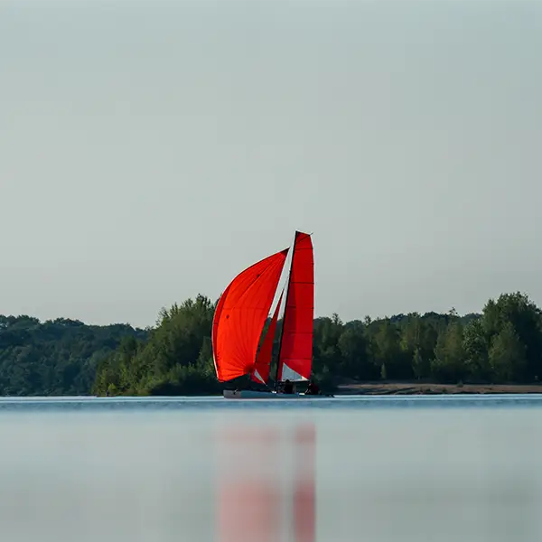Ein Segelboot mit leuchtend rotem Segel fährt auf einem ruhigen See, umgeben von bewaldetem Ufer.