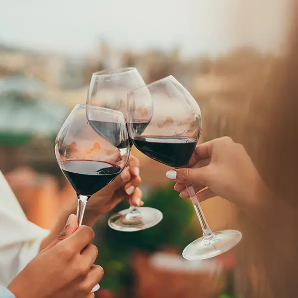 Drei Personen stoßen mit Gläsern Rotwein an, die im Freien bei weichem, warmem Licht gehalten werden.