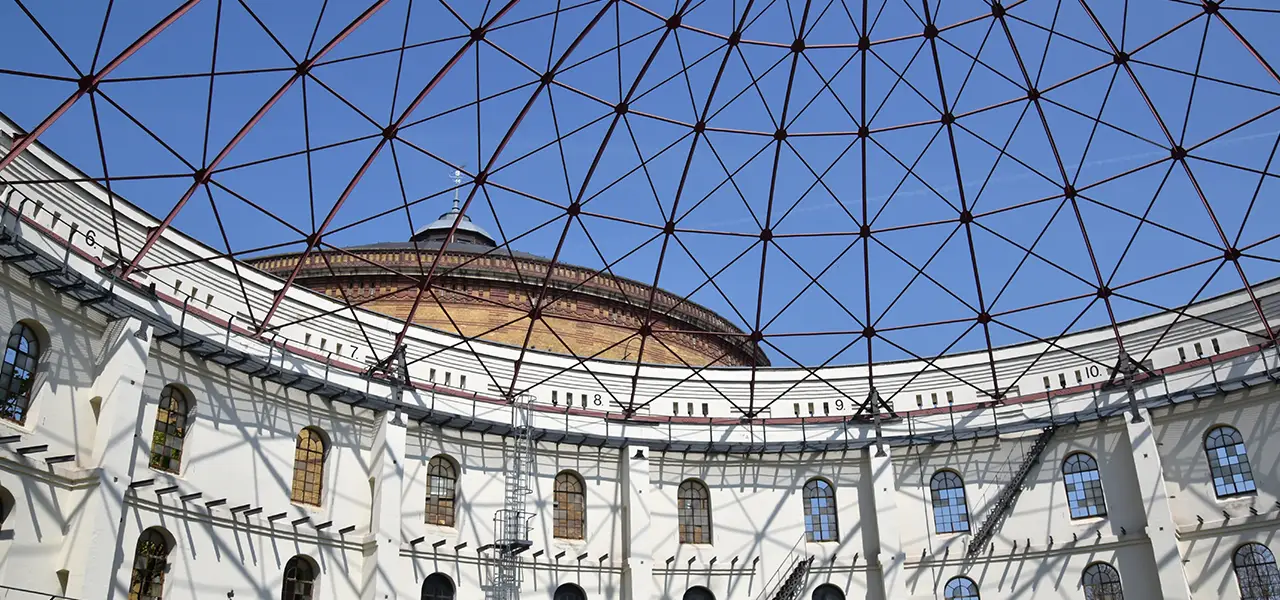 Blick auf das Innere des Panometer Leipzig mit einem kuppelförmigen Metallgerüst, das die runde Struktur überspannt. Unten sind weiße Wände mit hohen Fenstern zu sehen.