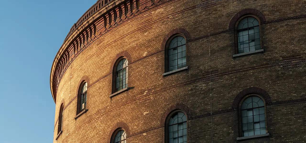 Nahaufnahme einer runden Backsteinfassade mit mehreren Reihen von großen, bogenförmigen Fenstern, die von Sonnenlicht beleuchtet werden.