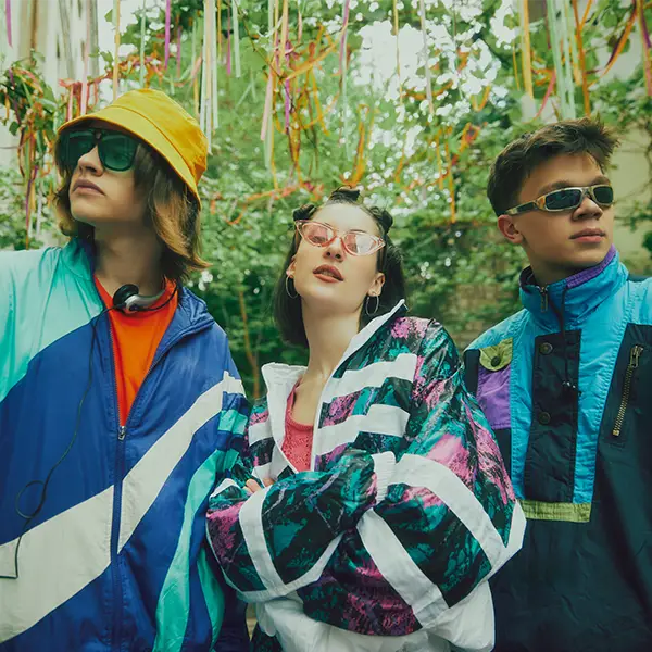 Drei junge Personen in bunten Retro-Trainingsjacken und Sonnenbrillen posieren in einem mit bunten Bändern dekorierten Outdoor-Bereich.