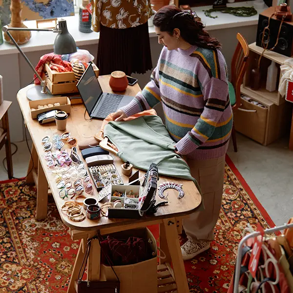 Eine Person im gestreiften Pullover arbeitet an einem Holztisch, der mit Stoffen, Accessoires und Nähmaterialien bedeckt ist. Ein Laptop steht auf dem Tisch, und der Raum ist mit bunten Teppichen und dekorativen Gegenständen eingerichtet.