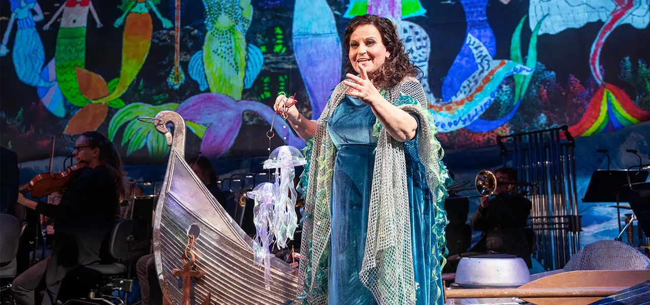 Frau in einem blauen Kleid auf einer Bühnenaufführung mit Meeresmotiv, mit einer Requisite eines Schiffes und einem farbenfrohen Hintergrund mit Meerjungfrauen.