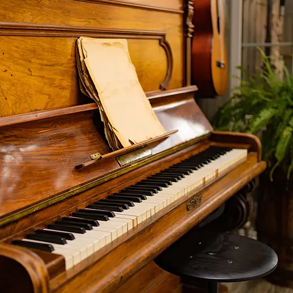 Ein detailreiches, antikes Klavier mit einer offenen Klaviatur, darauf alte, vergilbte Notenblätter und im Hintergrund eine akustische Gitarre und eine grüne Pflanze.