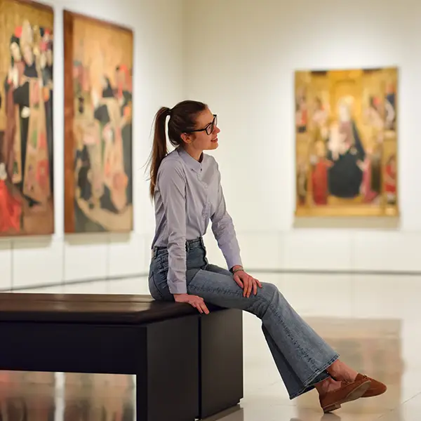 Eine junge Frau sitzt auf einer Bank in einem Kunstmuseum und betrachtet lächelnd die ausgestellten Gemälde an den Wänden.