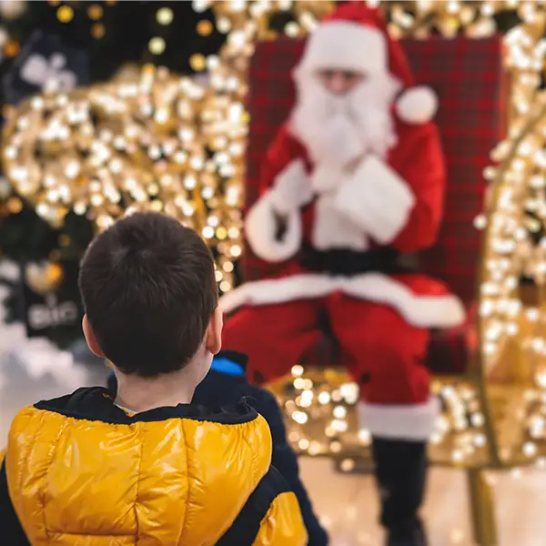 Ein Kind in einer gelben Jacke steht vor einem Weihnachtsmann, der auf einem mit Lichtern geschmückten Stuhl sitzt.